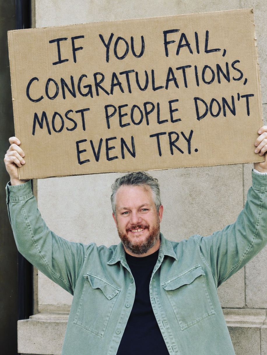 Rossco Paddison holding a cardboard sign reading 'If you fail, congratulations, most people don't even try.'